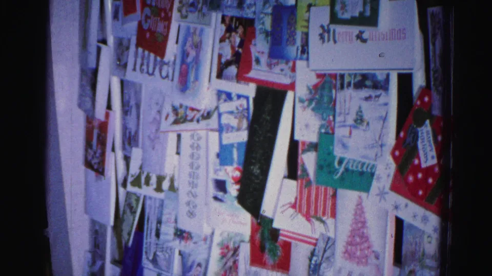Lower portion of a Christmas card wall display, 8mm home movie circa 1962 Hagerstown Maryland; pink Christmas tree card and 'Glad' text partially visible.