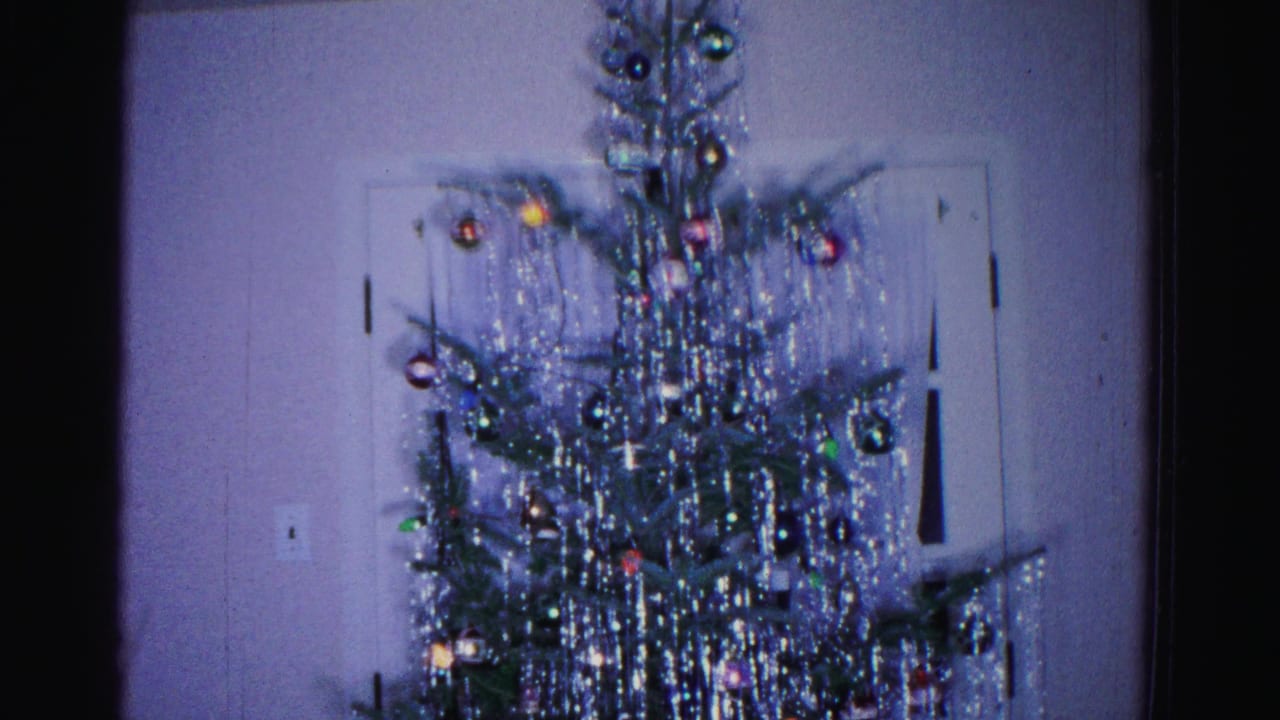 Upper portion of a heavily tinsel-draped Christmas tree with colorful ornaments, plain wall and white door behind.
