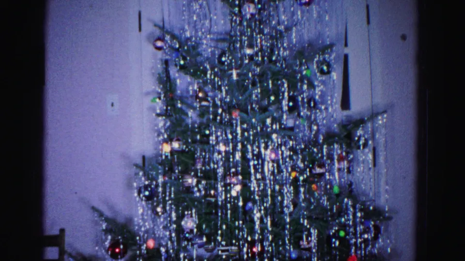 Lower section of a tinsel-laden Christmas tree filmed on 8mm, wooden chair beginning to appear in corner of domestic interior, circa 1962.