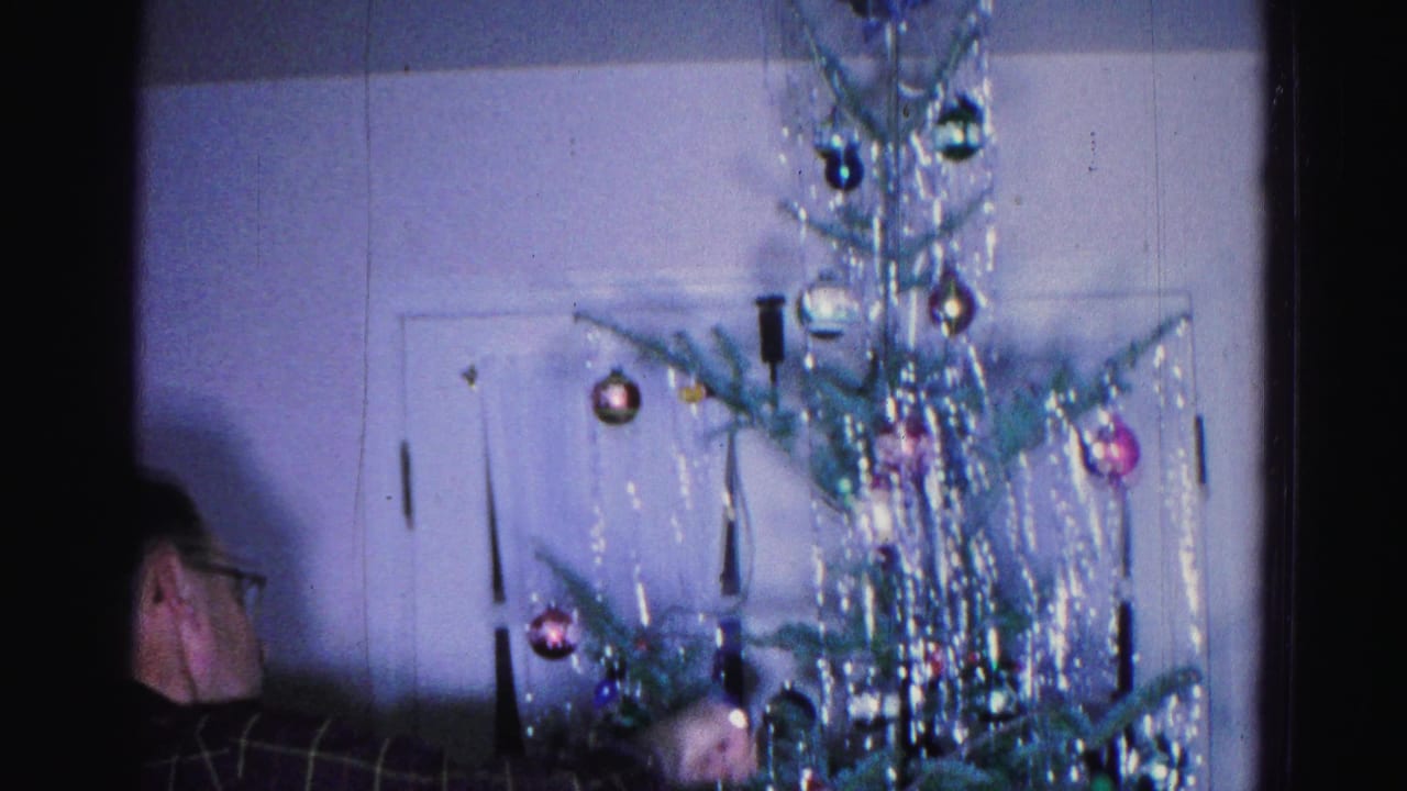Christmas tree with tinsel and colored ornaments centered in frame; partial profile of a man with glasses at left, white door in background.