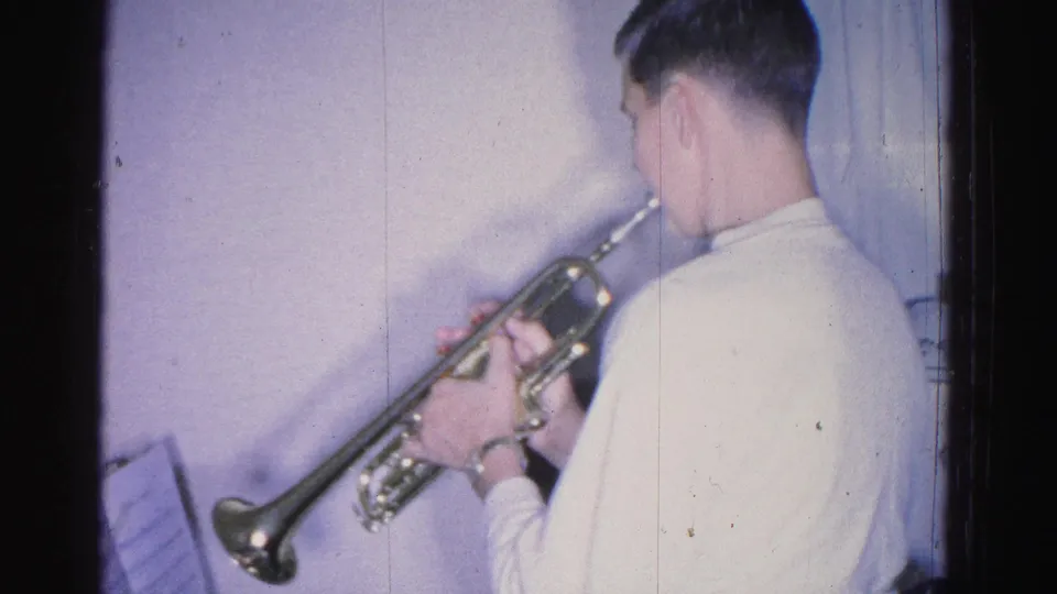 Young person in white shirt playing trumpet, seen from behind, with sheet music at frame edge; 8mm home movie interior, Hagerstown, Maryland, circa 1962.