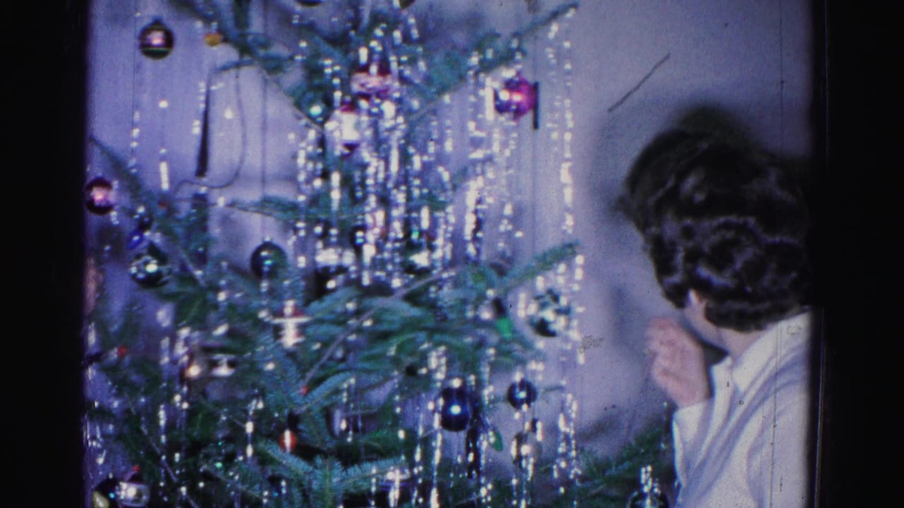A dark-haired adult stands to the right of a Christmas tree decorated with glass ball ornaments and heavy silver tinsel.