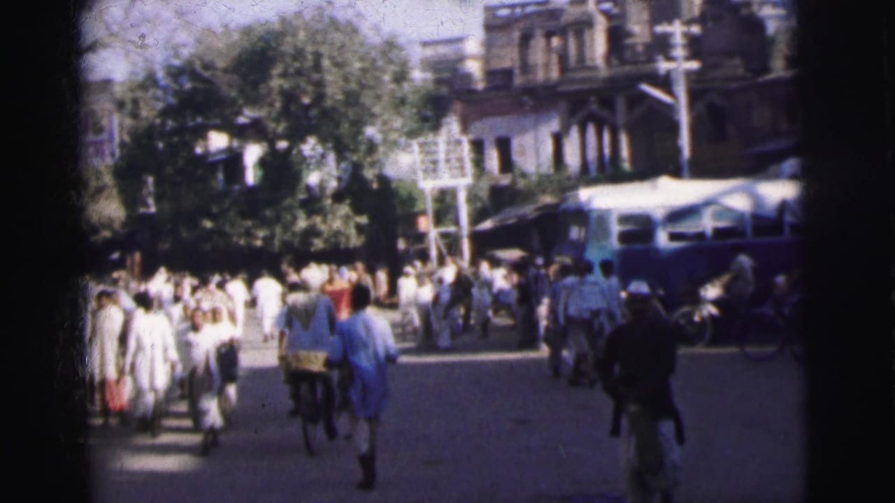 8mm home-movie frame from 1962 Calcutta showing pedestrians in white clothing on a busy street lined with shade trees and colonial-era buildings.