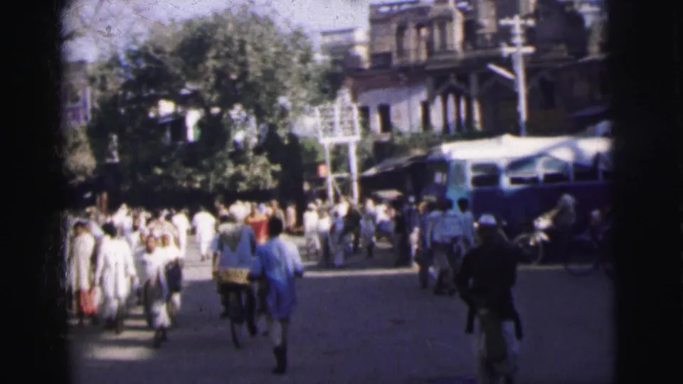 8mm home-movie frame from 1962 Calcutta showing pedestrians in white clothing on a busy street lined with shade trees and colonial-era buildings.