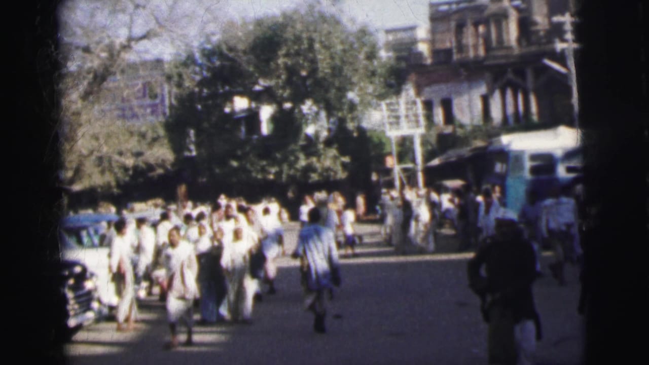 8mm footage from 1962 Calcutta showing pedestrians in traditional dress approaching the camera, with a dark period sedan and blue bus visible among shade trees and colonial buildings.