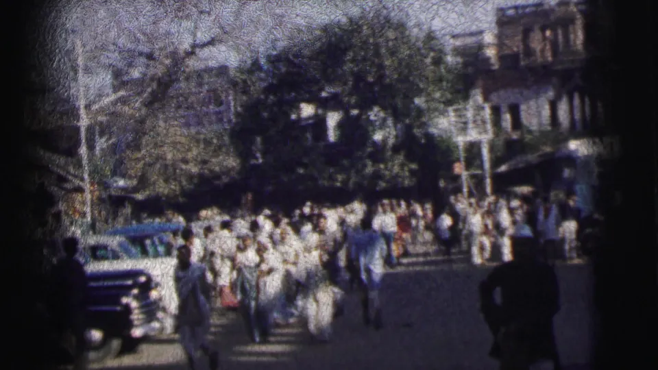 8mm frame from 1962 Calcutta showing a dense crowd filling a wide street flanked by large trees and colonial-era multi-story buildings.
