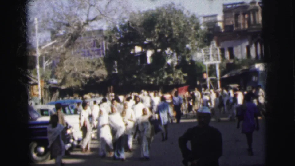 Final 8mm frame from 1962 Calcutta showing a moving crowd in white clothing, dark period cars on the right, and a tree-lined colonial streetscape.