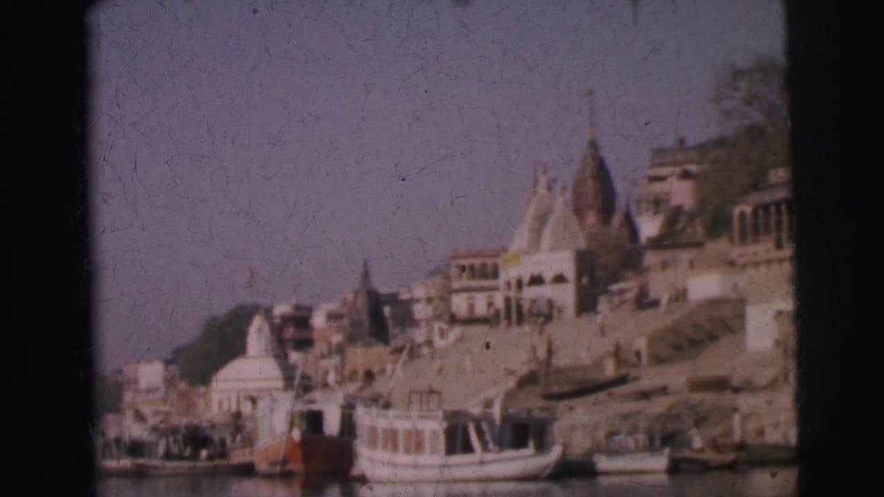 8mm home movie frame, circa 1962, showing Hindu temple shikhara towers and tiered ghat buildings rising above an Indian river, with white boats moored at the bank.