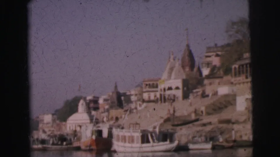 8mm home movie frame, circa 1962, showing Hindu temple shikhara towers and tiered ghat buildings rising above an Indian river, with white boats moored at the bank.