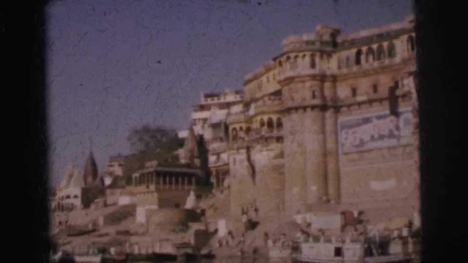 8mm frame, circa 1962, showing ornate multi-story ghat buildings with arched balconies, curved temple towers, and trees along an Indian riverfront.