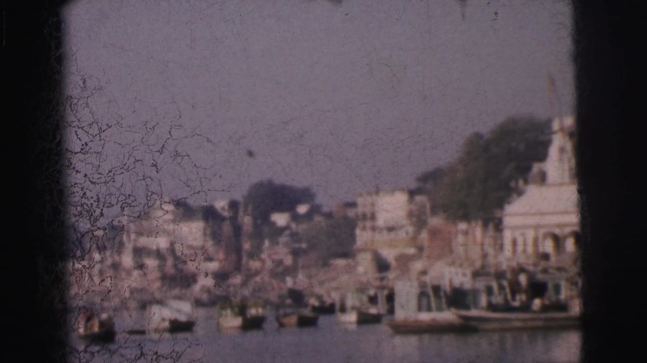 Wide river shot with white-domed temples and buildings on the far bank, wooden boats moored in the foreground, heavy 8mm film grain and emulsion crackle visible.