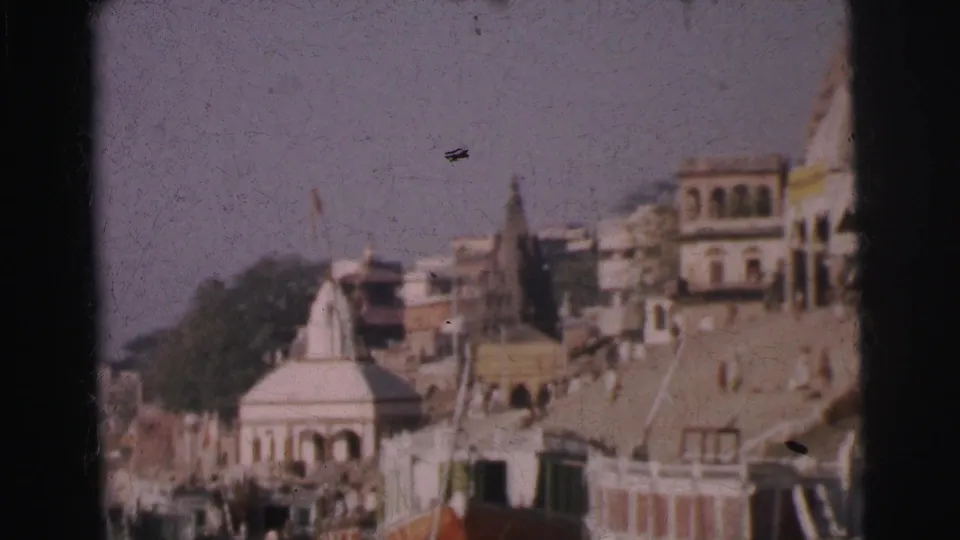 8mm colour film, India circa 1962: white temple spire and coloured riverside buildings above a stepped embankment, moored boats below, possible pennant visible.