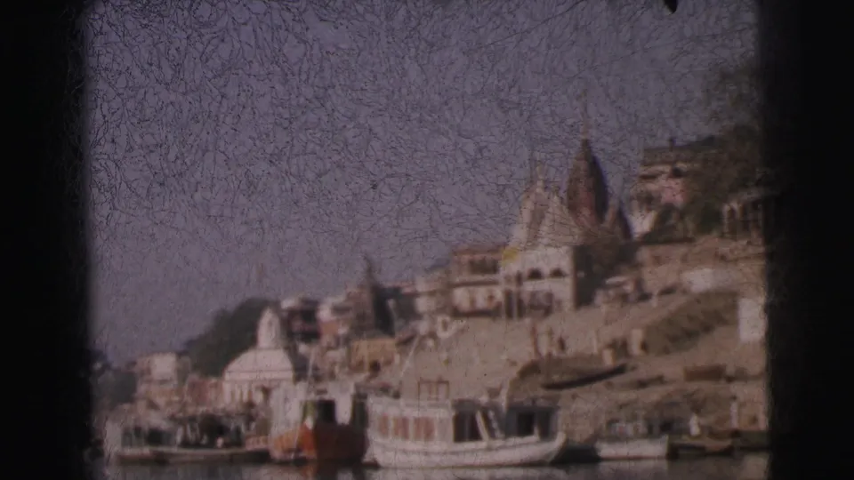 8mm colour footage, India circa 1962: pointed temple shikharas and stacked riverside buildings on a sloped embankment, small boats moored at the water's edge.