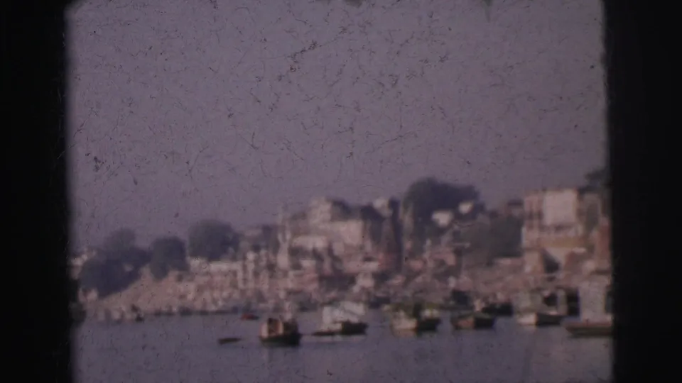 8mm home-movie frame circa 1962 showing Calcutta's densely built riverfront with several small boats moored at the bank and brownish building tones.