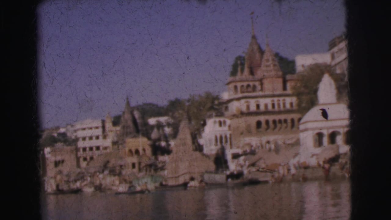 Temple shikharas and white dome above river ghat steps, small figures visible at waterline, 8mm film.