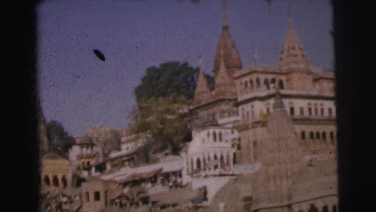 Establishing 8mm frame of a stepped ghat with red-stone temple spires, whitewashed multi-story buildings, market stalls below, and blue sky, India, circa 1962.
