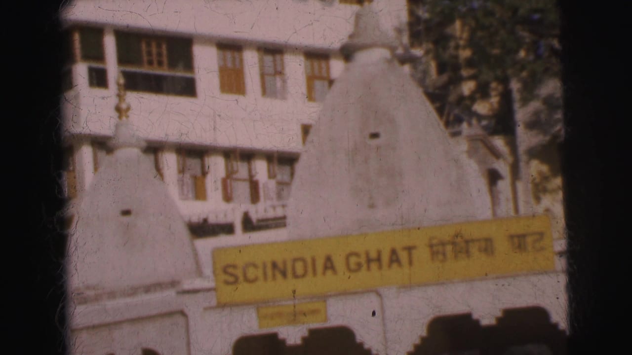 Close 8mm frame of yellow enamel bilingual sign reading SCINDIA GHAT in English and Devanagari script, mounted on whitewashed building, small white dome shrine in foreground, circa 1962.