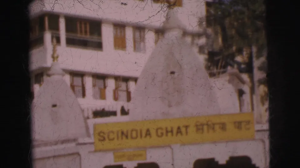 8mm frame showing yellow bilingual Scindia Ghat sign at lower edge, whitewashed multi-story building with wooden-shuttered windows and small white dome, India, circa 1962.