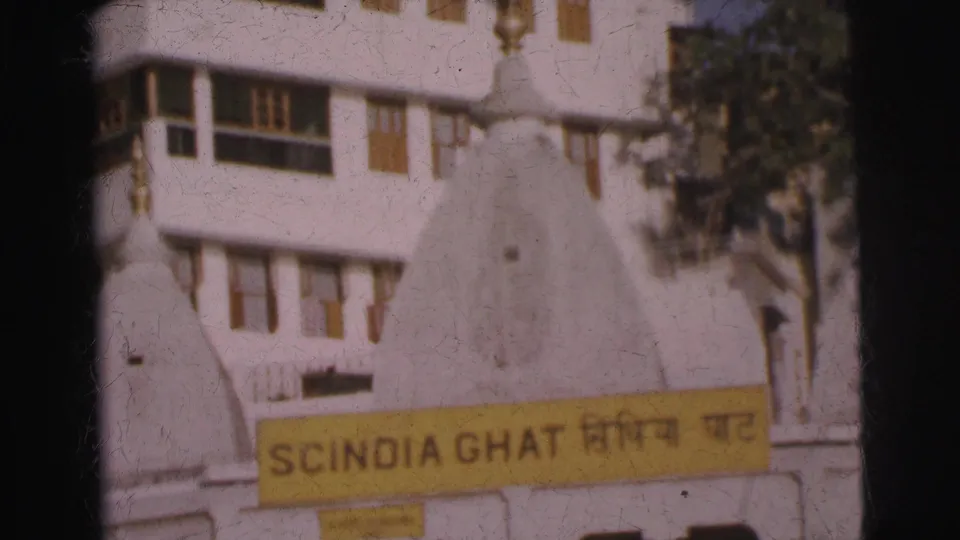 8mm frame of yellow bilingual Scindia Ghat sign on whitewashed building with a small white dome and green trees at right, India, circa 1962.
