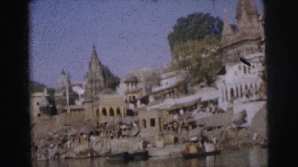 8mm colour frame, c. 1962, of a busy Indian riverfront showing multi-storey colonnaded buildings, terracotta temple spires, ghat steps, and small wooden boats.
