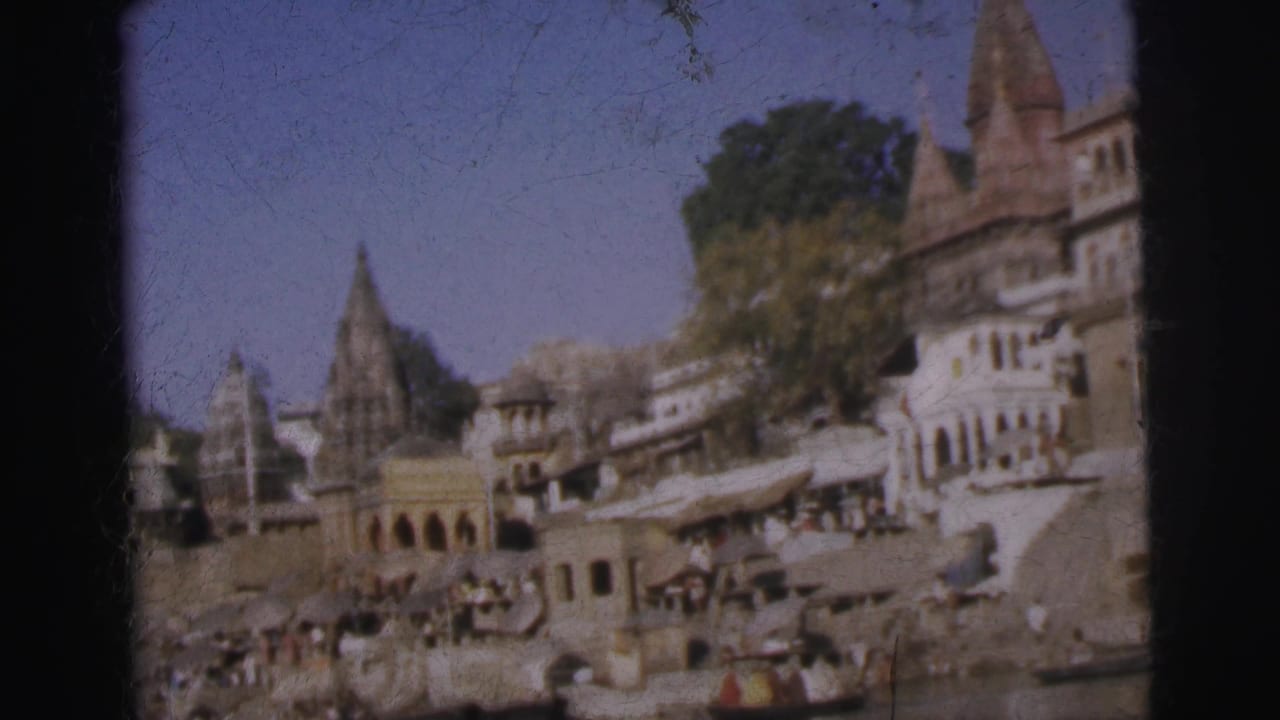 Slightly shifted river-level view of the same Indian ghat, showing temple towers and multi-storey riverside buildings with people on the steps.
