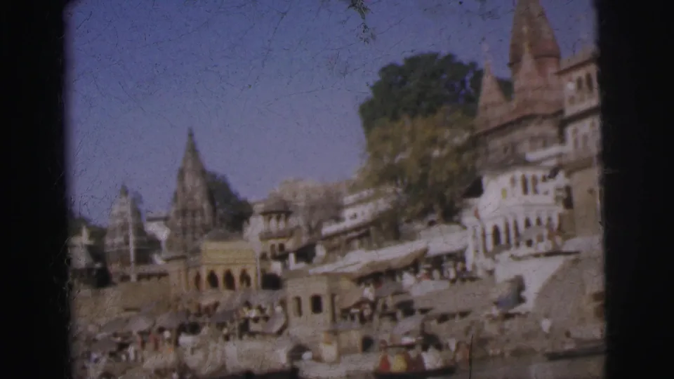 8mm colour frame, c. 1962, panning slightly right along an Indian riverfront ghat with temple spires, arched colonnades, and people gathered on the steps.