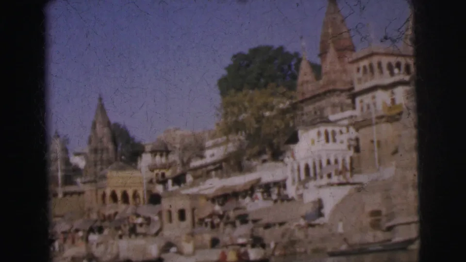 8mm colour frame, c. 1962, tightened on prominent temple towers and riverside buildings of an Indian ghat, with people and market awnings visible at water level.