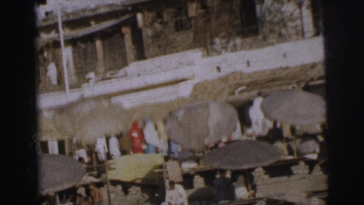 Similar ghat scene with crowd on steps under umbrellas; slight camera shift reveals more of the upper stone building facade.