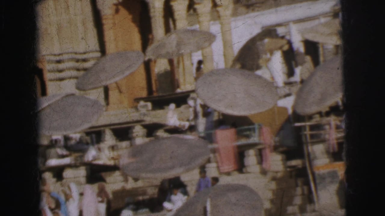Overhead shot of a crowded market with large round thatched parasols and figures in white garments on stone pathways.