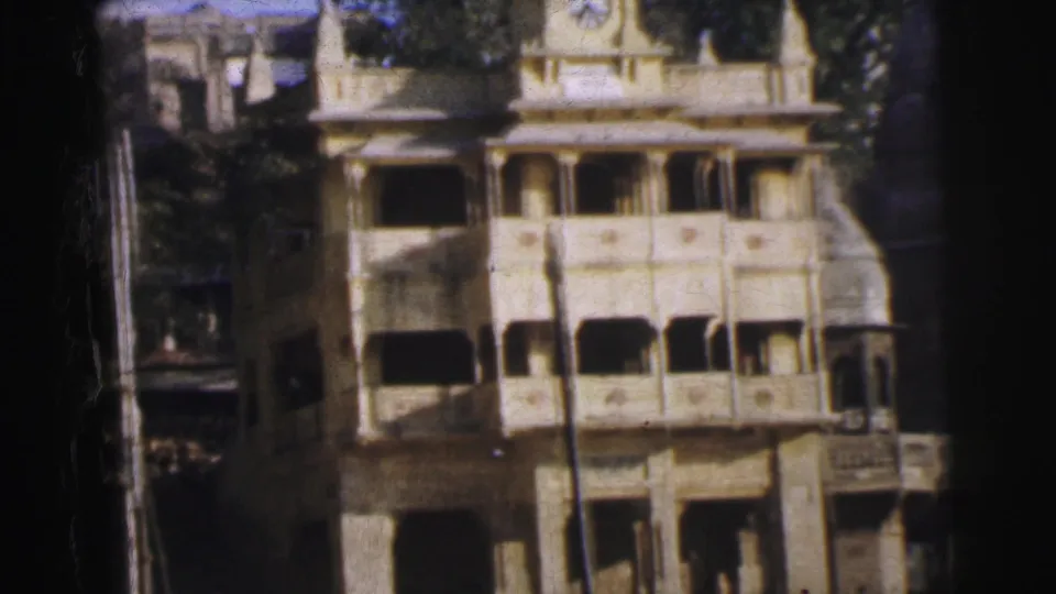 Multi-story colonial-style building in Calcutta, circa 1962, showing tiered arched balconies, a wooden pole, and trees on aged 8mm color film with warm color shift.