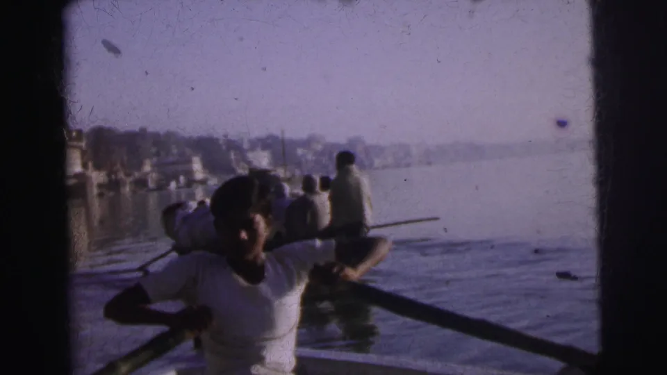 A young man rows a wooden boat on the river at Calcutta, circa 1962, with passengers behind him and a hazy distant cityscape along the far bank.