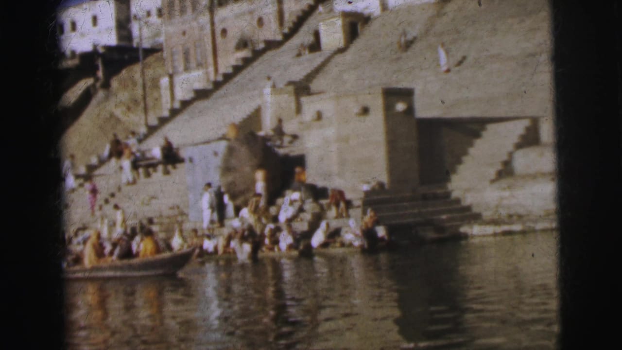 Wide shot of a stone river ghat with a crowd gathered at the water's edge and a small wooden rowboat.