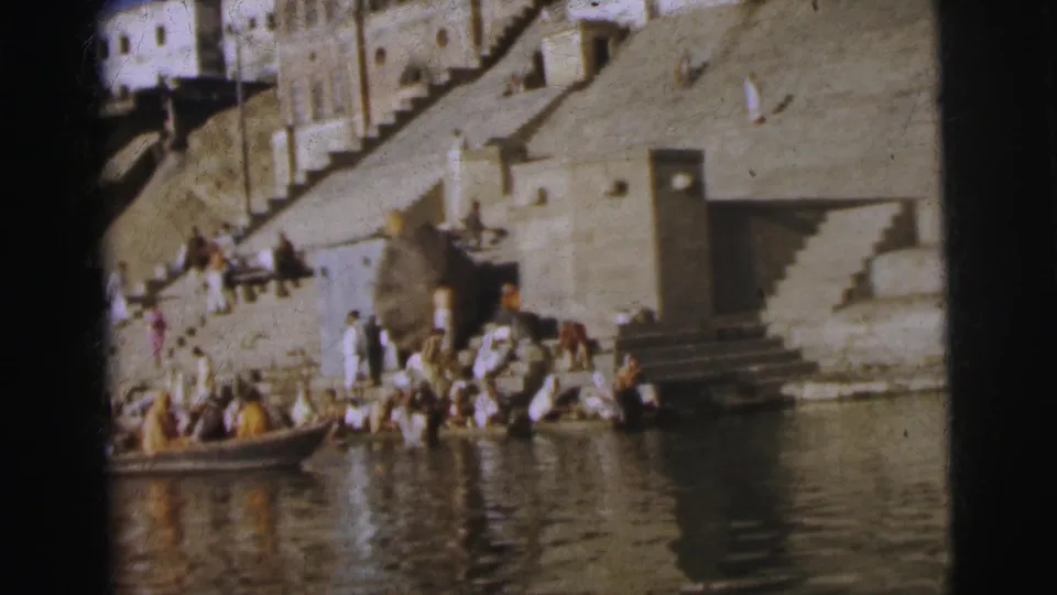 Wide 8mm shot of a stone ghat in Calcutta, 1962, showing a crowd at the waterfront with a wooden rowboat alongside the steps.