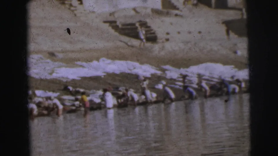 8mm home movie frame showing communal laundry washing at a stone ghat in Calcutta, 1962, with white fabric prominently spread on the bank.