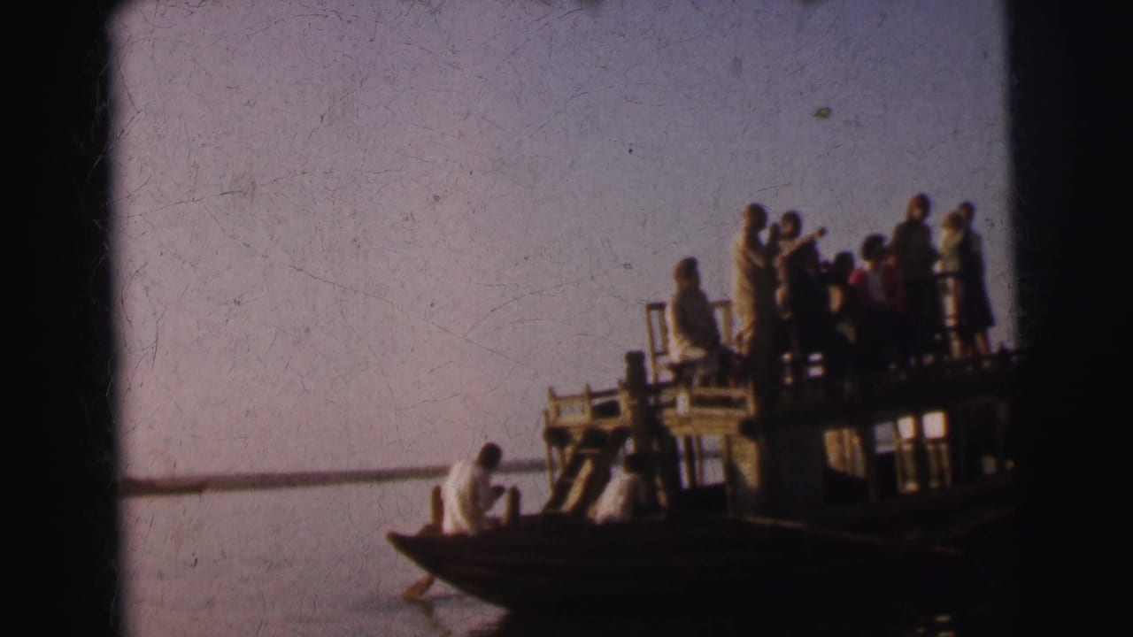 Group of people standing on an elevated wooden platform barge on a river, with a man in white in a smaller boat alongside.