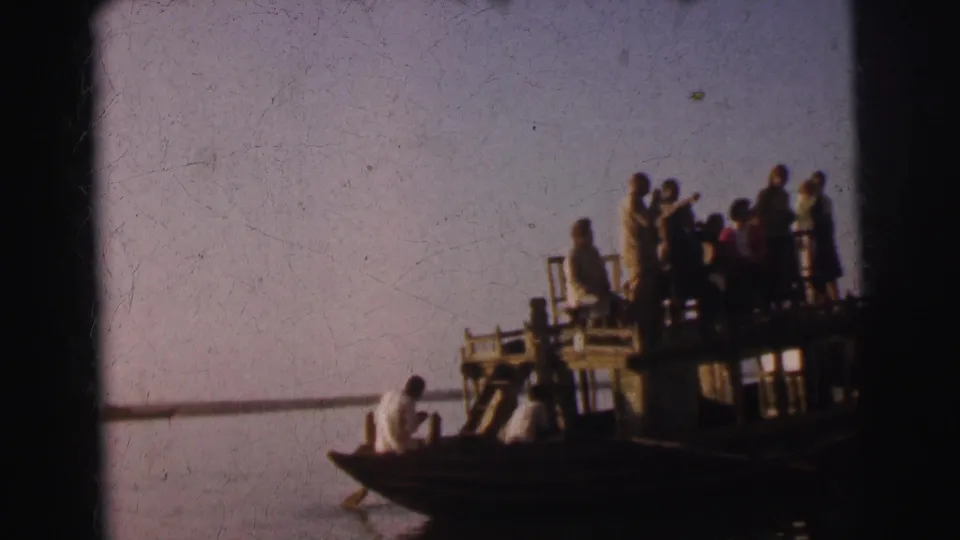 Group of roughly ten figures in period dress standing on a raised wooden boat platform on a calm river in Calcutta, India, circa 1962, shot on 8mm film.