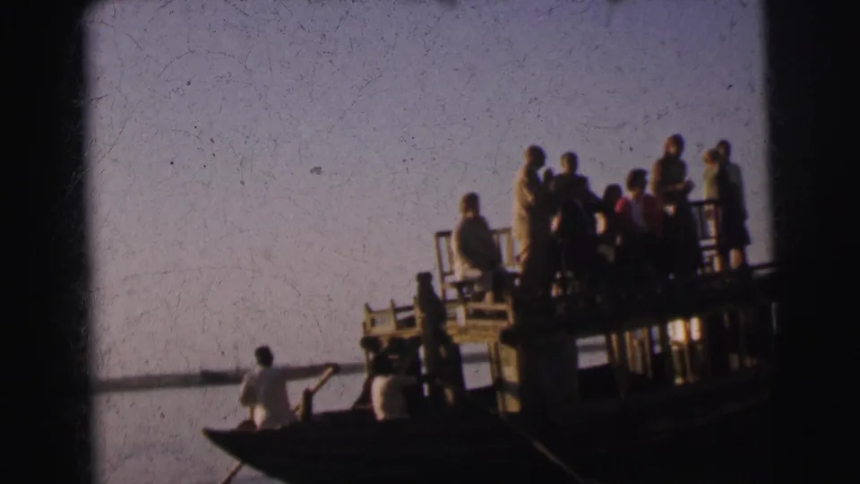 Group of figures standing on a raised platform boat on a calm river, with a white-shirted oarsman in an adjacent rowboat, circa 1962 Calcutta, India.