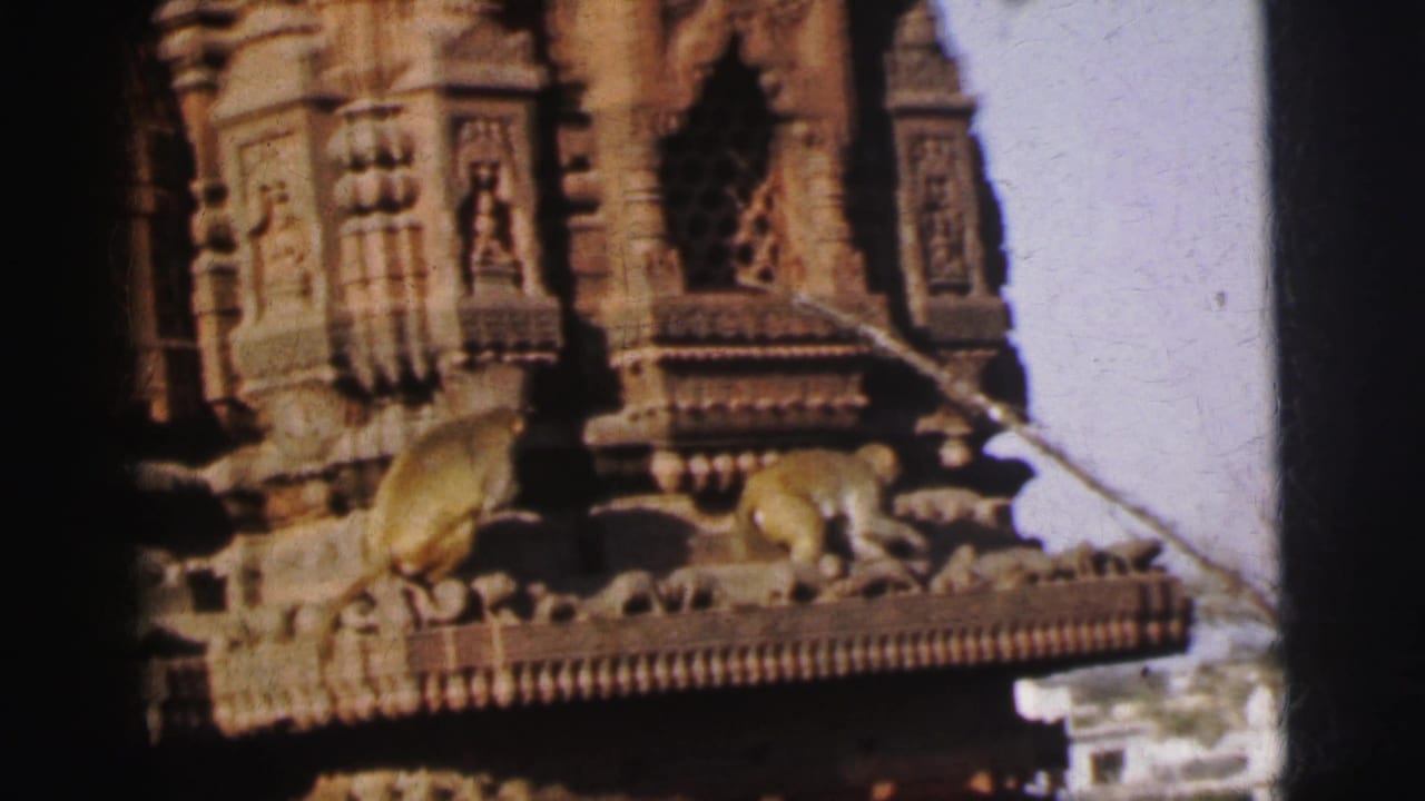Two or three small primates walk along the carved stone cornice ledge of an ornate temple tower under blue sky.