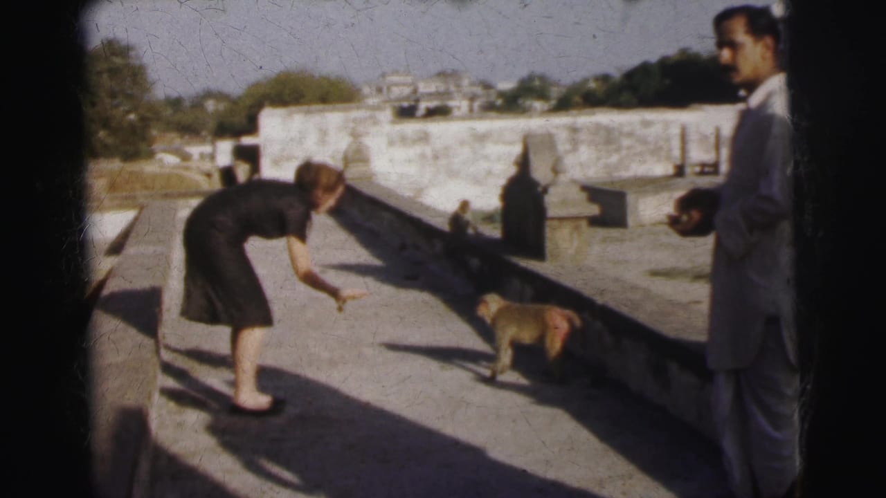 Western woman leaning toward a macaque on a temple rooftop parapet in what appears to be 1960s India; a man in a dark jacket observes from the right.