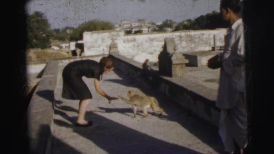 Woman crouching to interact with a macaque at terrace-step level on what appears to be a 1960s Indian temple rooftop; male figure stands in background.