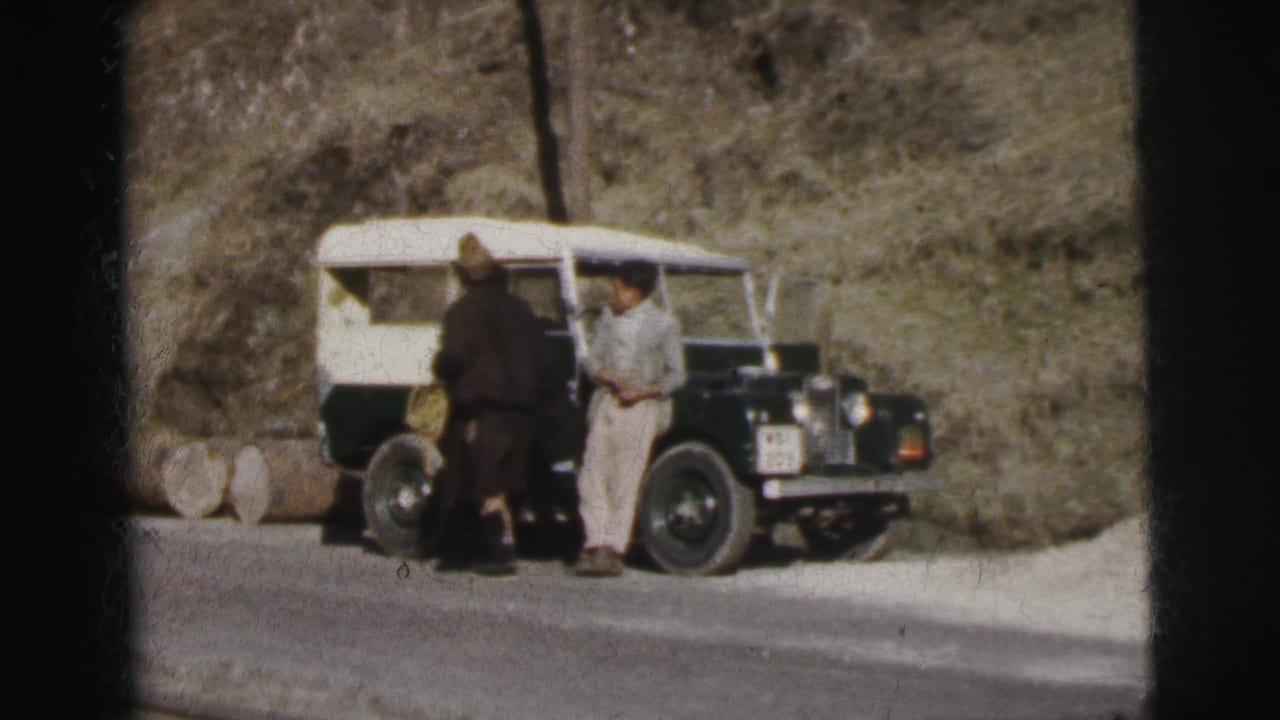 Two men standing beside a dark green and white Land Rover on a narrow mountain road, rocky hillside behind.