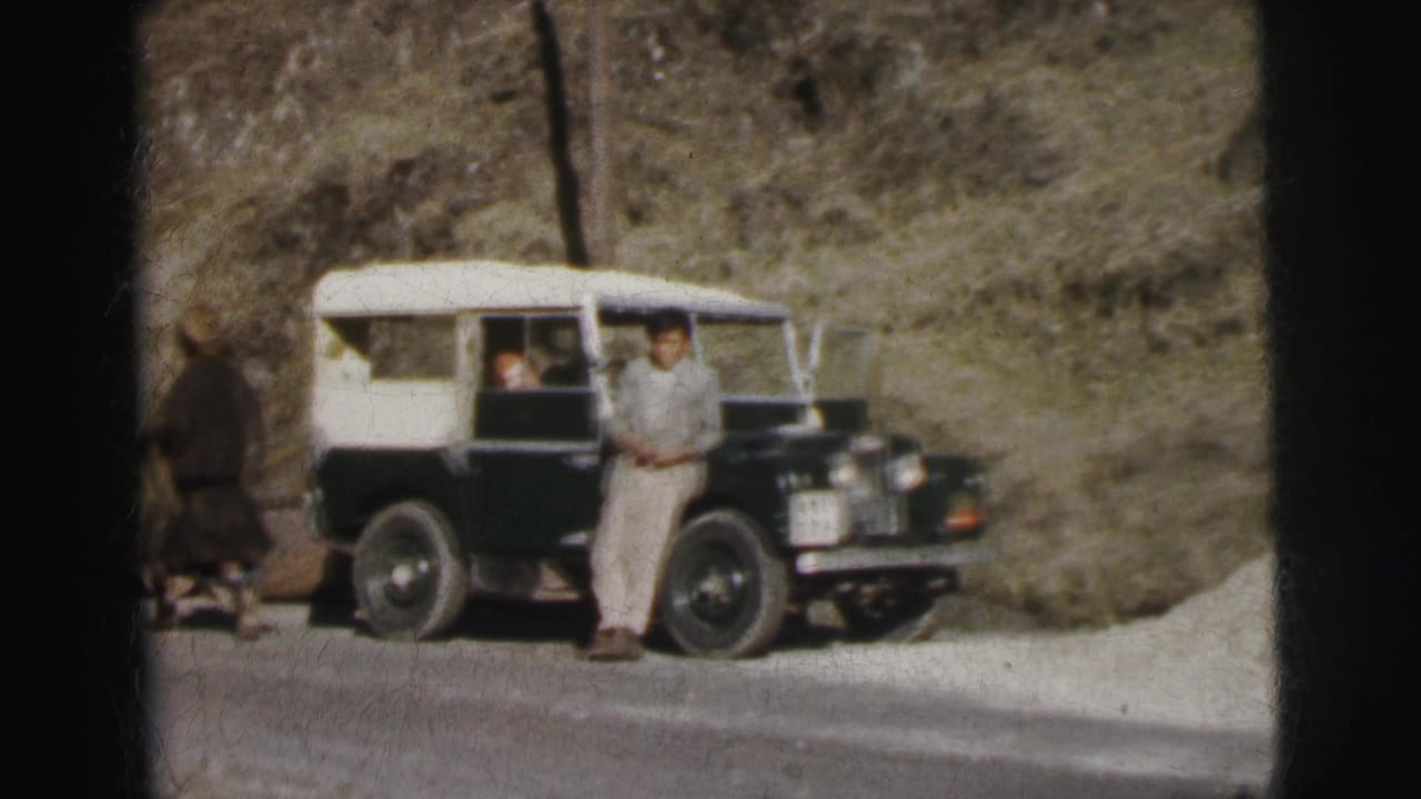Two men near a two-tone Land Rover on a mountain road; one figure turning away, one standing beside the vehicle.