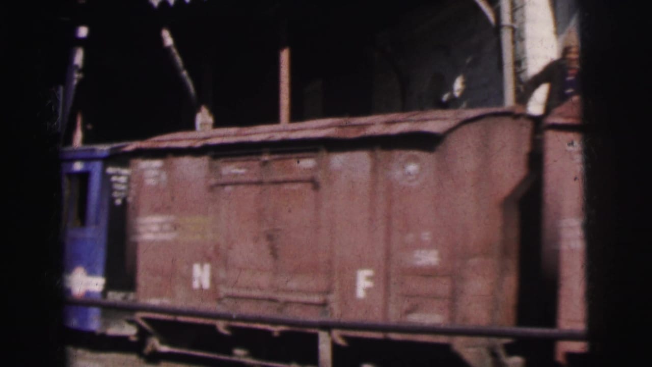Close shot of a weathered brown freight car marked 'N' and 'F' in white paint, with a blue railcar partially visible at left, at a railway yard.