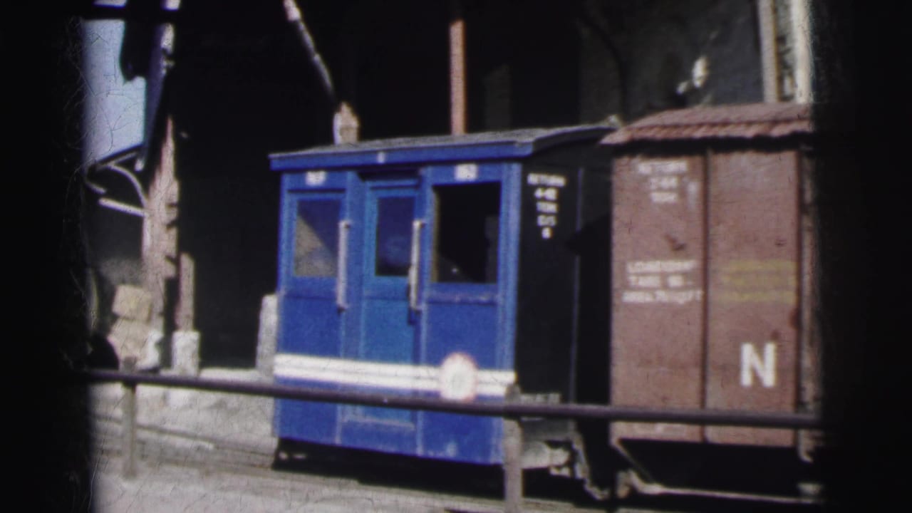 A compact blue railcar with white horizontal stripe and small windows stands at a rail yard, with a brown freight car marked 'N' at right.