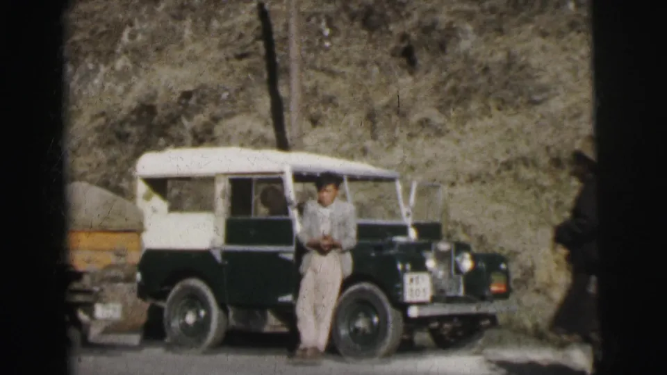 8mm film circa 1962, India: a man in pale trousers stands at the driver's door of a green and white Land Rover on a narrow mountain road, with a steep earthen embankment behind.