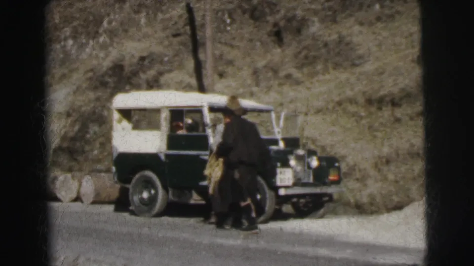 8mm film circa 1962, India: two people are visible near a green and white Land Rover on a narrow mountain road — one standing at the vehicle, one approaching in a dark coat or shawl.