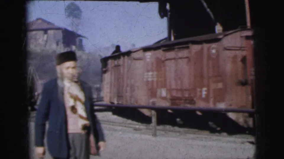 Man in traditional dark hat and blue jacket at a Calcutta railway yard, c.1962; rust-brown freight cars with white stenciled lettering behind him.