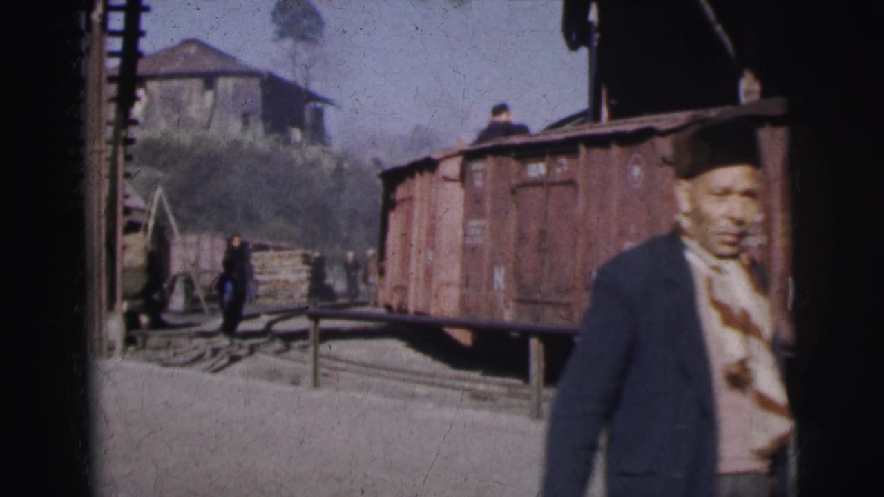 Man in dark hat facing right in foreground; a second person walks the yard and stacked timber is visible behind rust-red freight wagons.