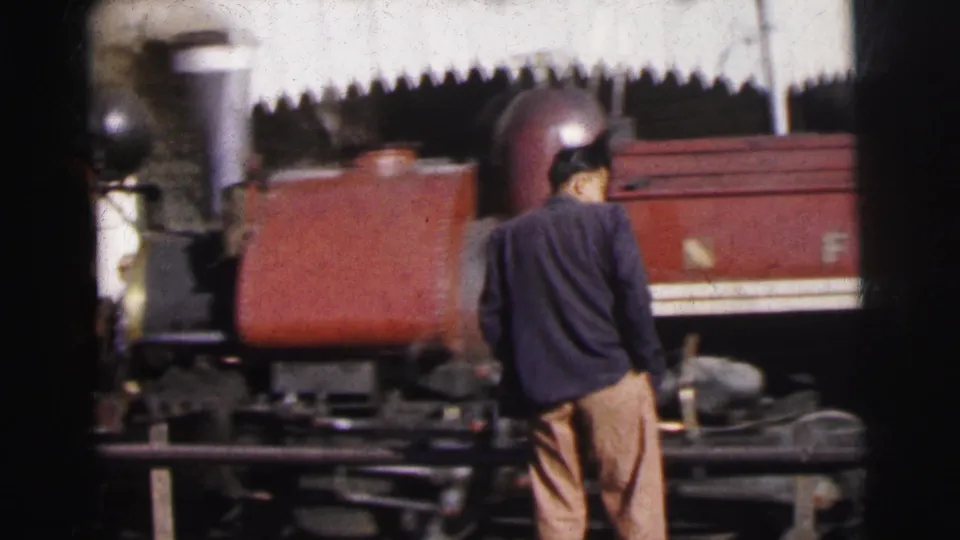 Adult male observer in dark jacket and light trousers stands trackside, back to camera, watching a rust-red steam locomotive, Calcutta railway yard, circa 1962, 8mm film.