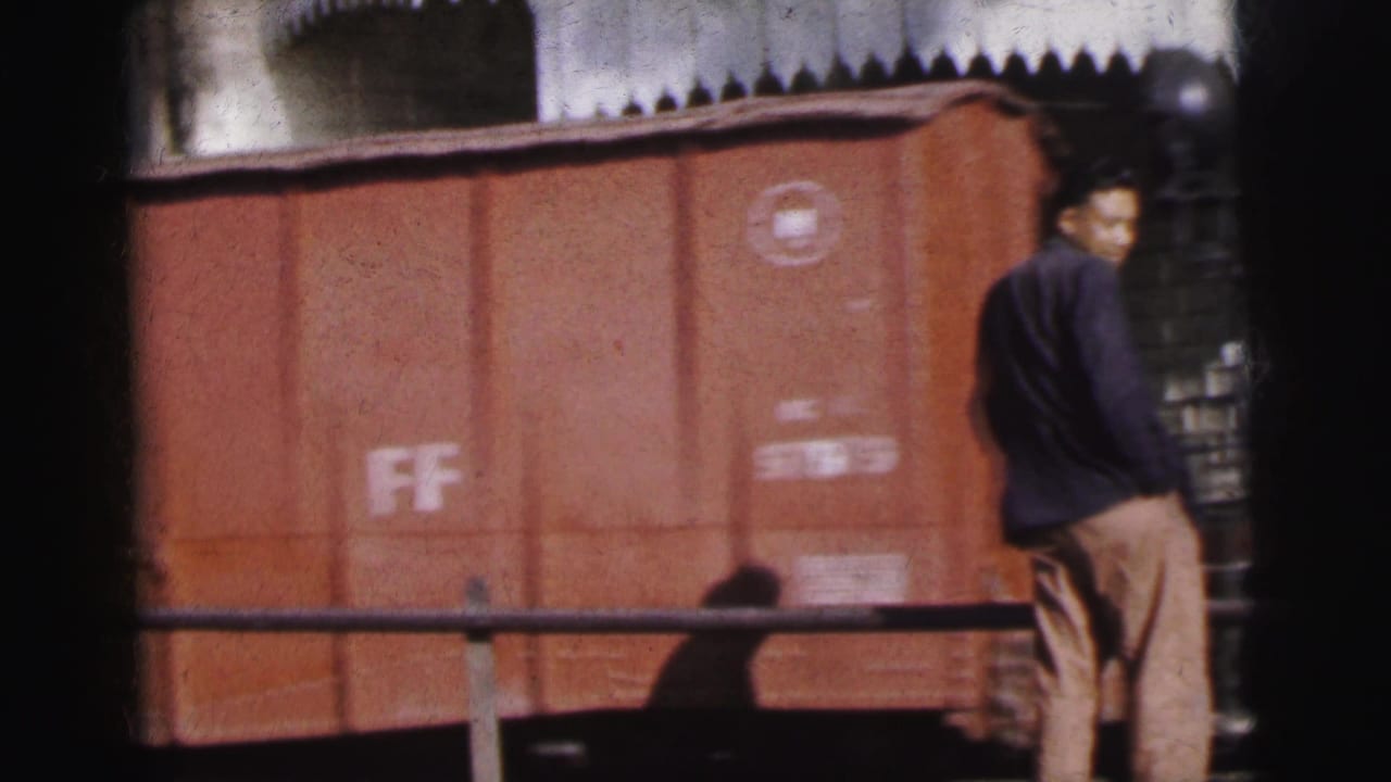Man in dark jacket stands in profile watching a large rust-red boxcar stencilled FF pass along the tracks.